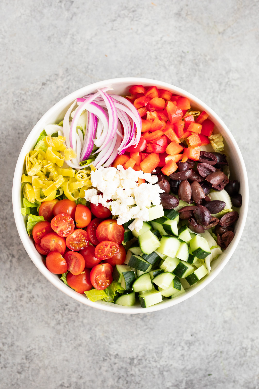 Italian chopped salad ingredients arranged in a bowl with romaine, cucumber, tomatoes, red pepper, olives, feta, red onion, and pepperoncini